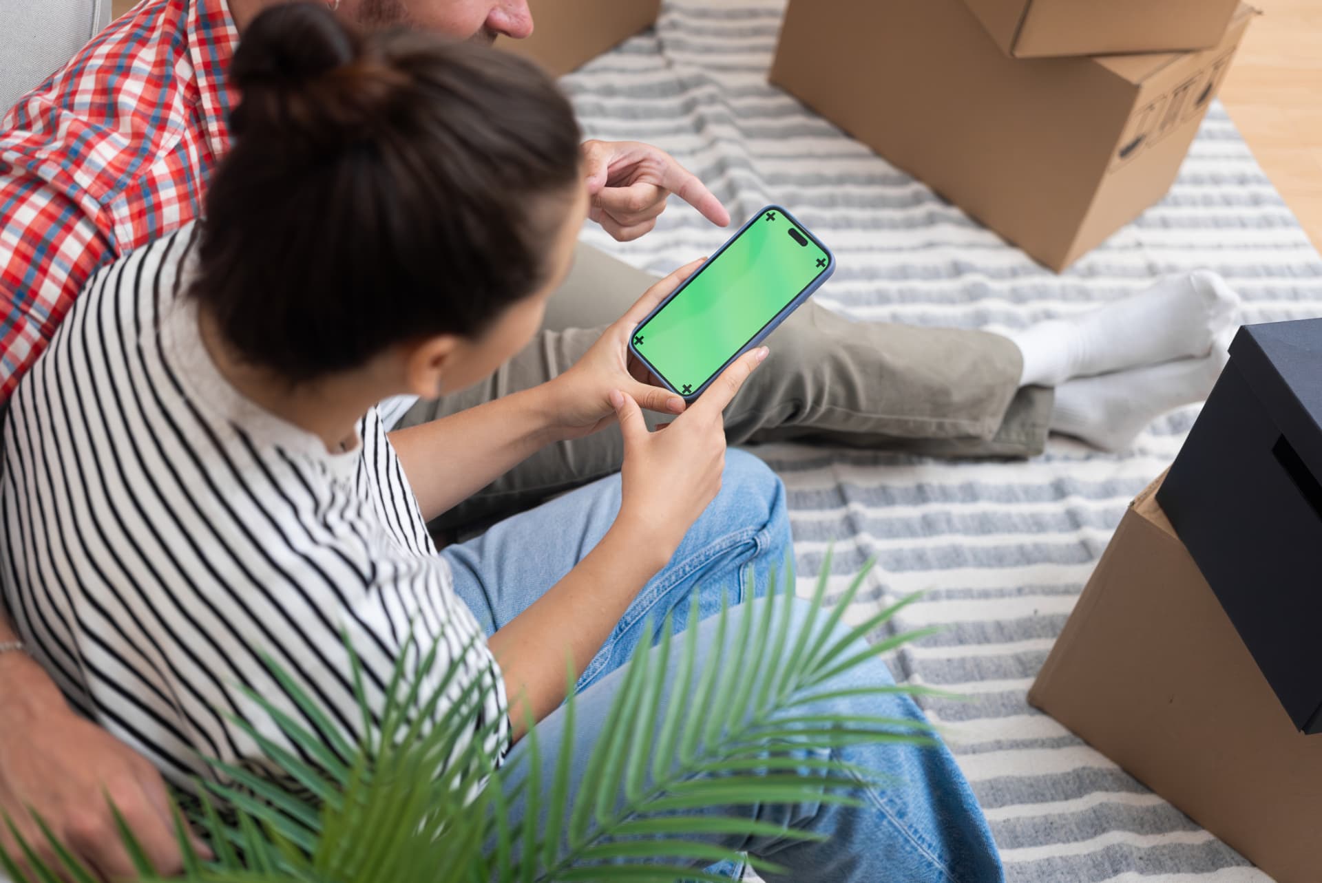 Couple assis parmi ses cartons d’emménagement, recherchant un logement sur smartphone