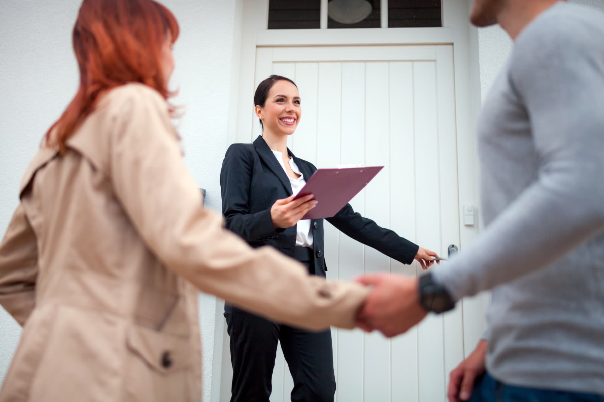 Une agente immobilière souriante, porte-bloc en main, ouvre la porte d’un logement à un couple qui se tient par la main
